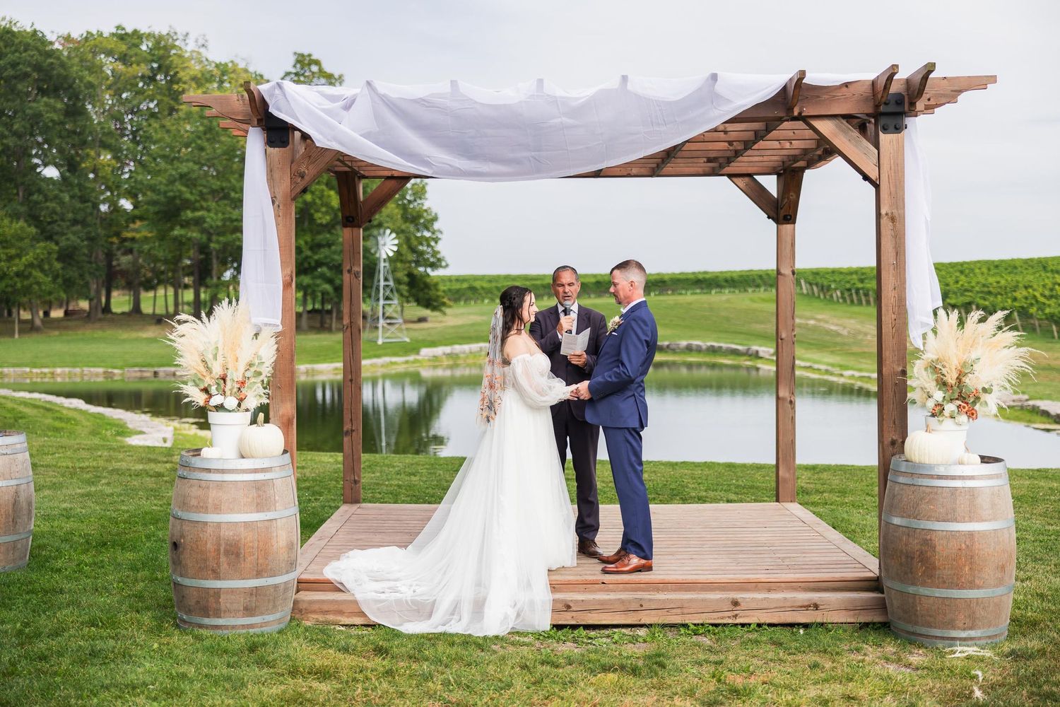 Couple stands under wooden pergola with draped fabric and wine barrels at outdoor wedding.