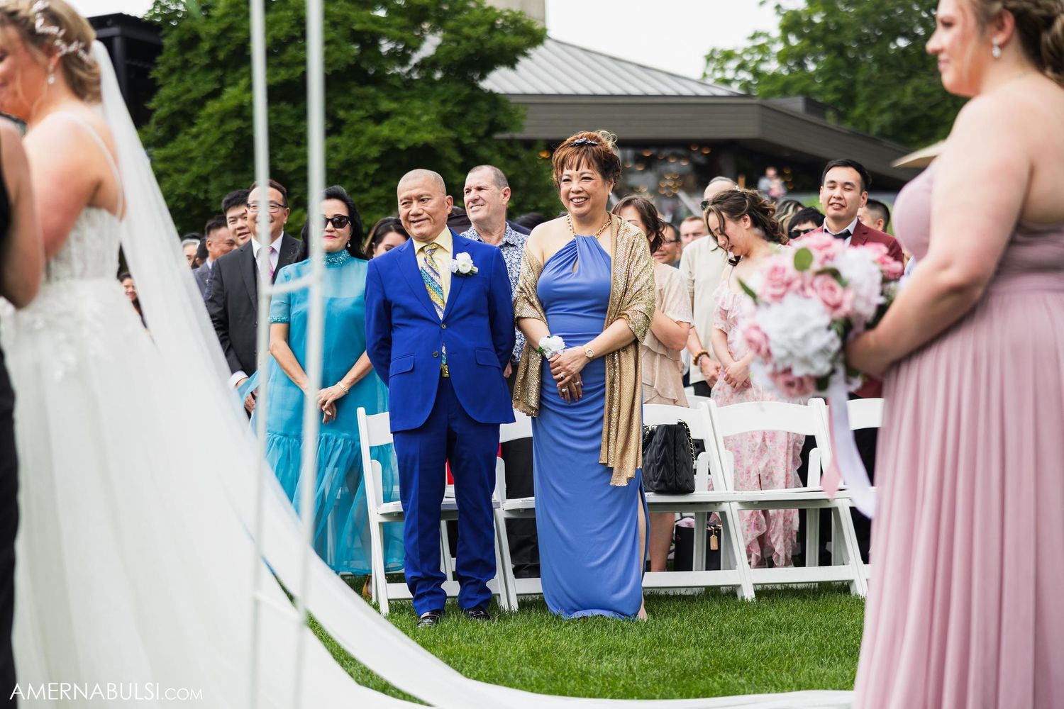 Wedding guests and attendants in formal attire gathered on a manicured lawn during an outdoor celebration.
