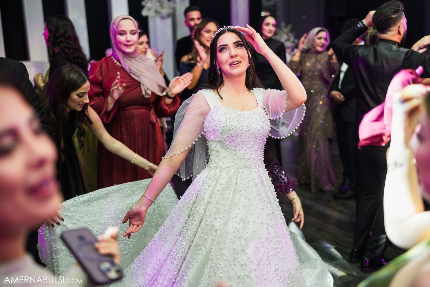 A bride in a white wedding gown dances joyfully surrounded by guests at a reception celebration.