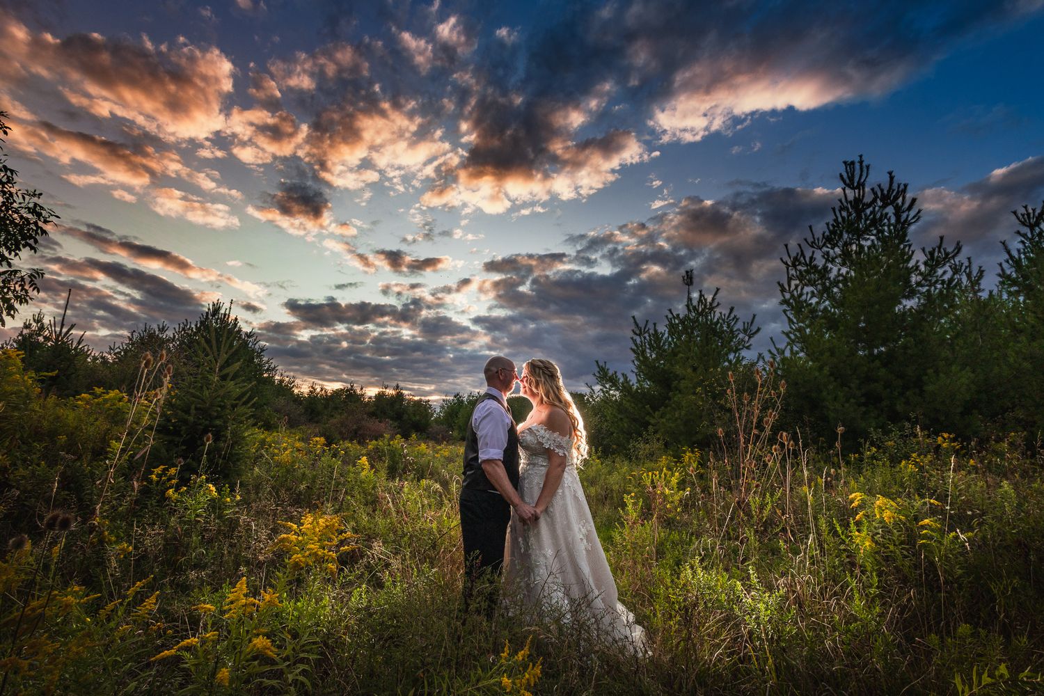 Wedding couple strolls through a field at golden hour with dramatic sunset clouds and desert landscape.