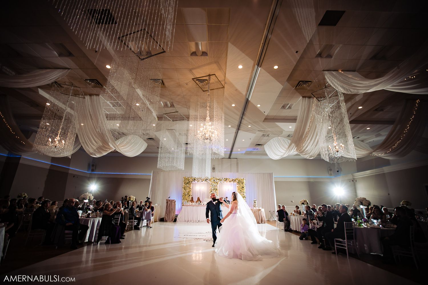 Wide panoramic view of an elegant white ballroom wedding reception with guests seated at round tables.