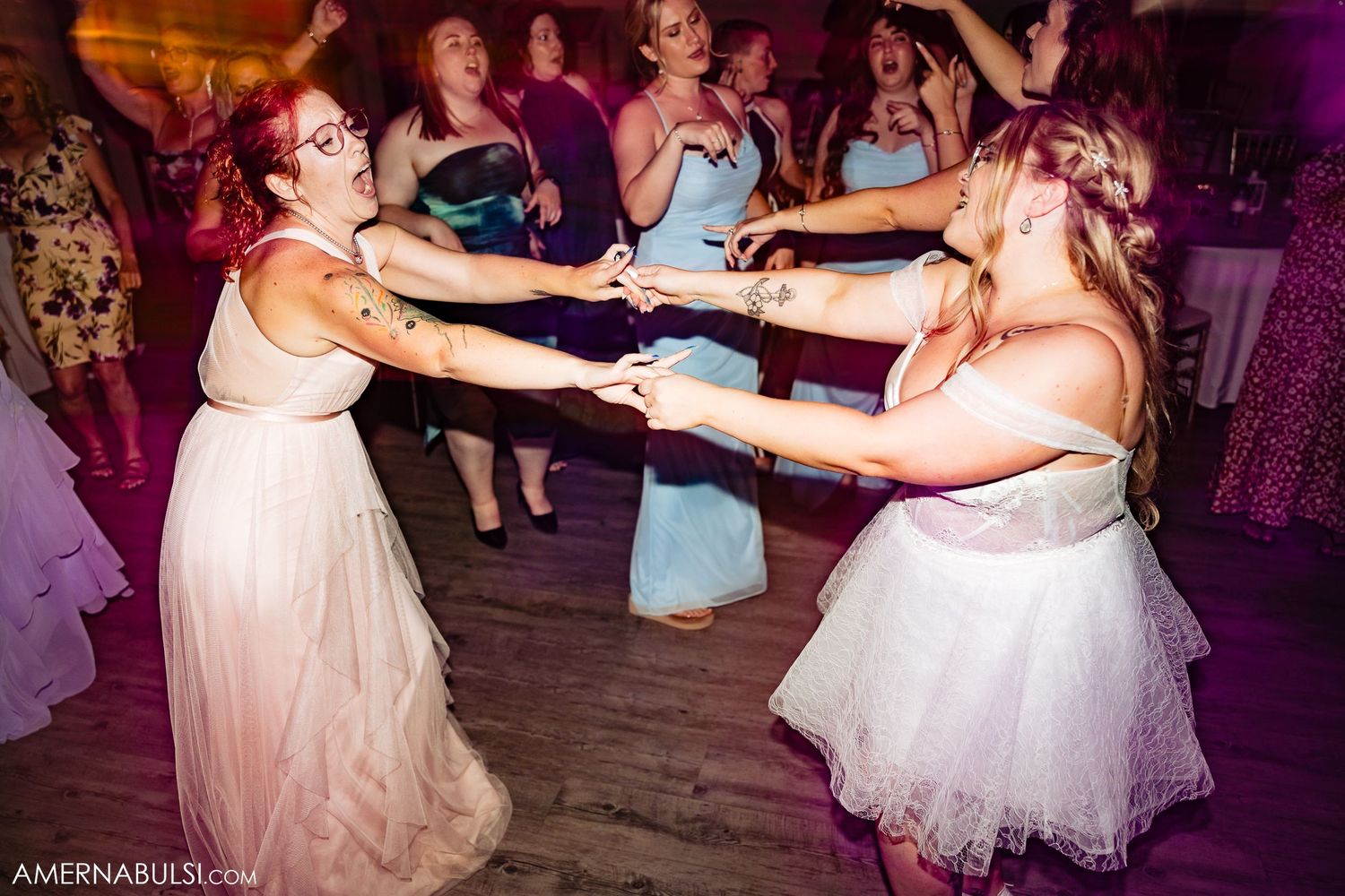 A group celebrates on the dance floor at a wedding reception, with dancers wearing blue and white formal dresses.