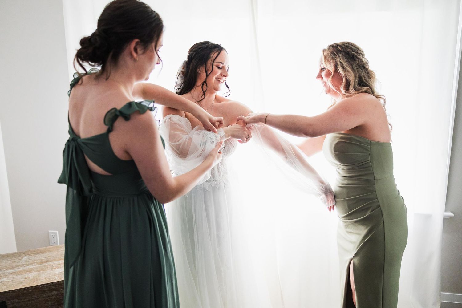 Bridesmaids in green dresses help the bride get ready in a bright room.