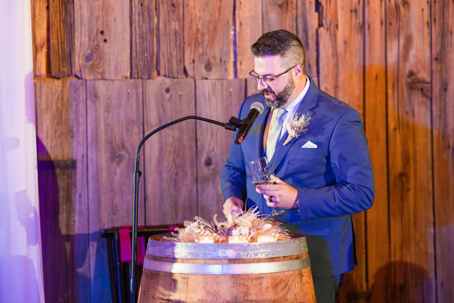 A speaker addresses wedding guests from behind a rustic wine barrel podium under purple lighting.