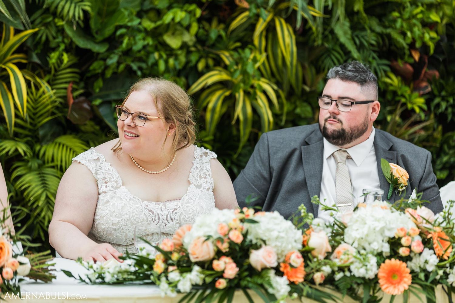 A couple sits at their wedding reception table decorated with peach and white floral arrangements.