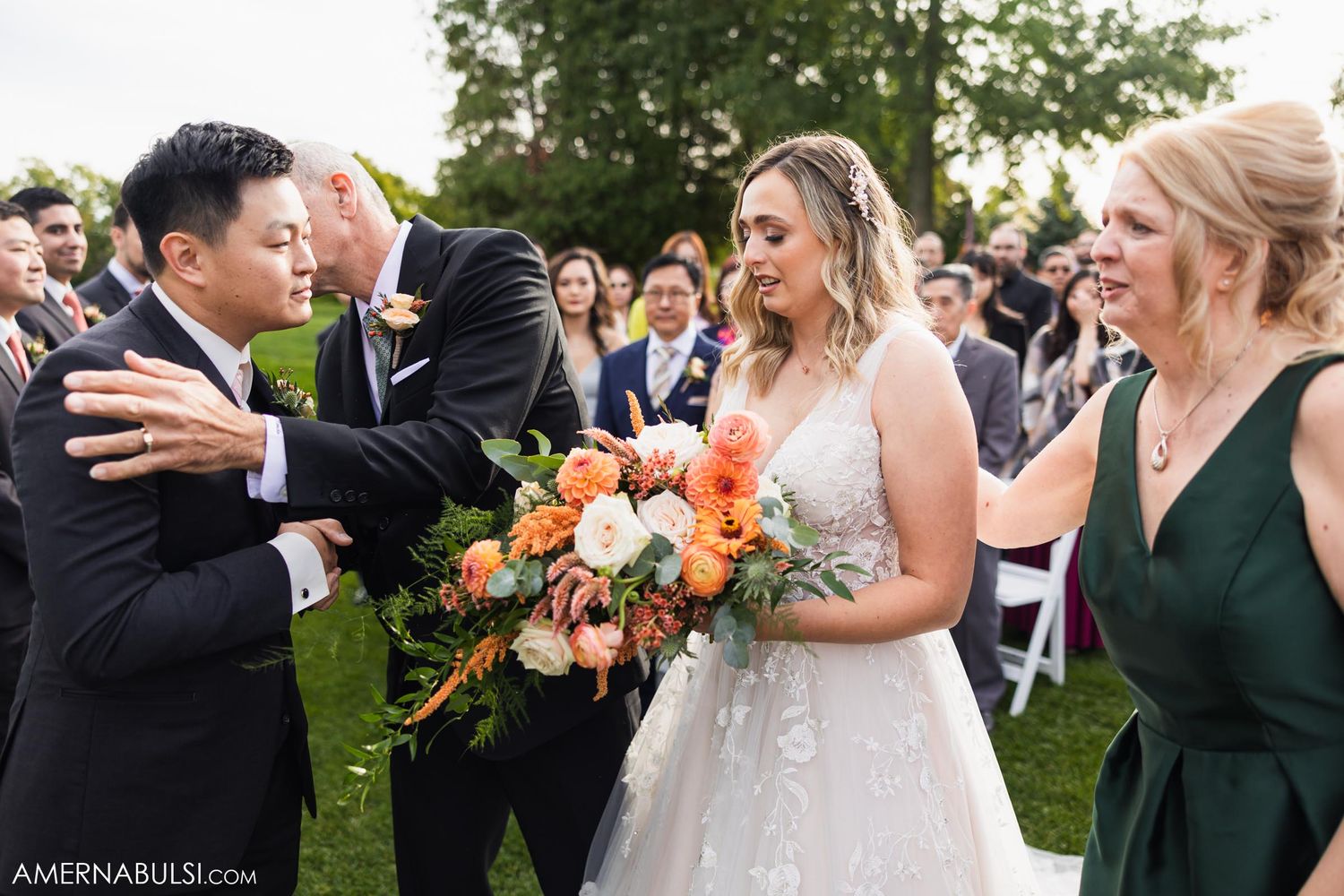 Wedding party sharing emotional moment during outdoor ceremony with orange floral bouquet.