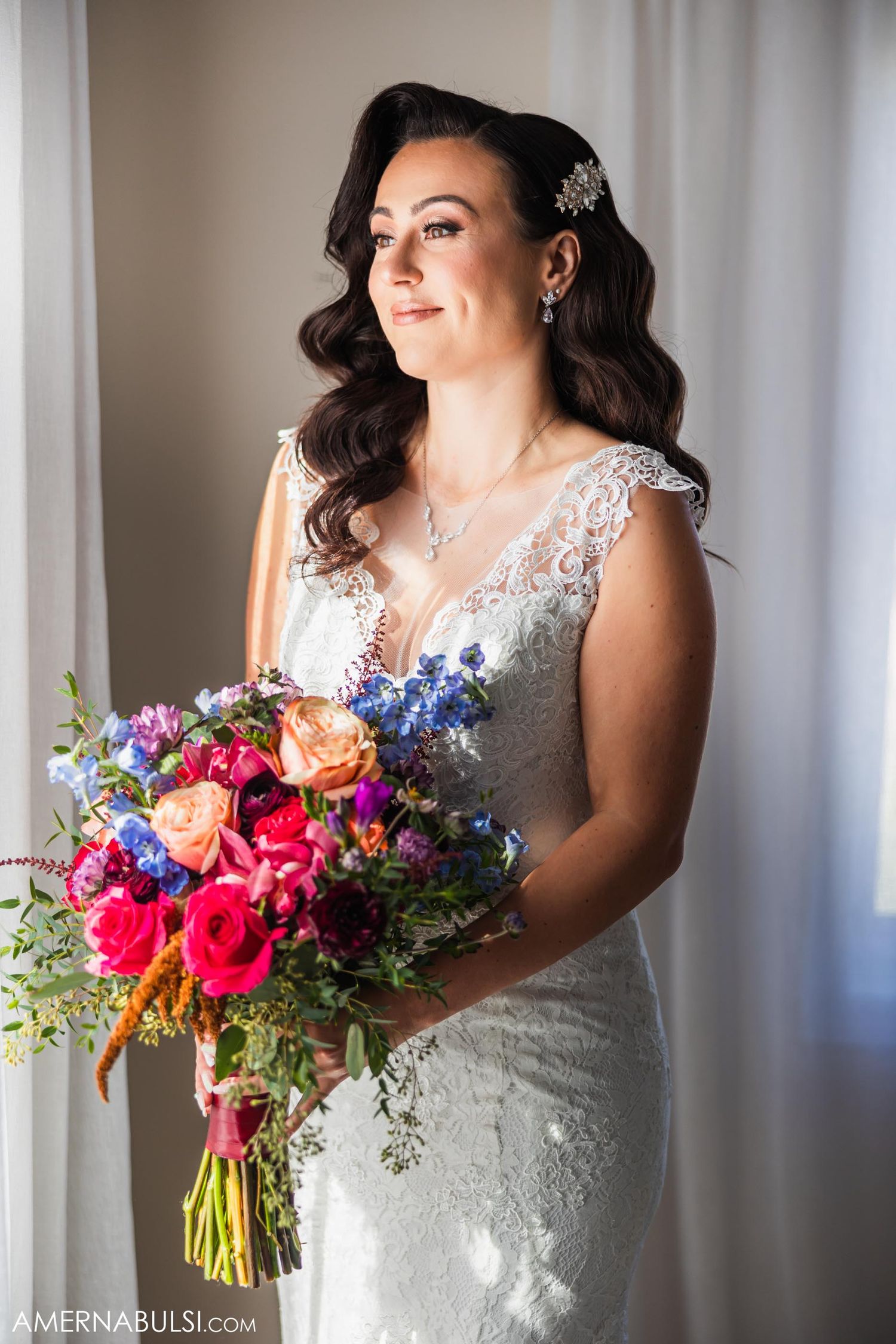 A bride in a lace wedding dress holds a vibrant bouquet of pink, blue and orange flowers.