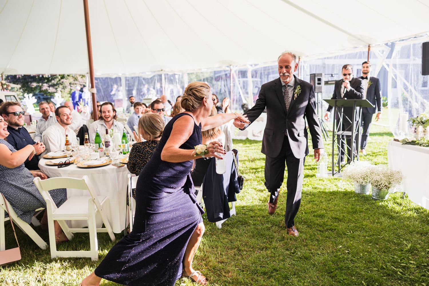 Wedding guests dancing and celebrating under white tent during daytime reception.