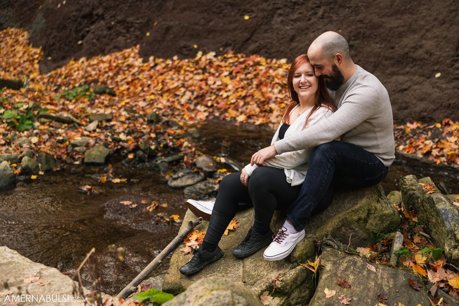 Couple sits together on rocky ledge surrounded by fallen autumn leaves.