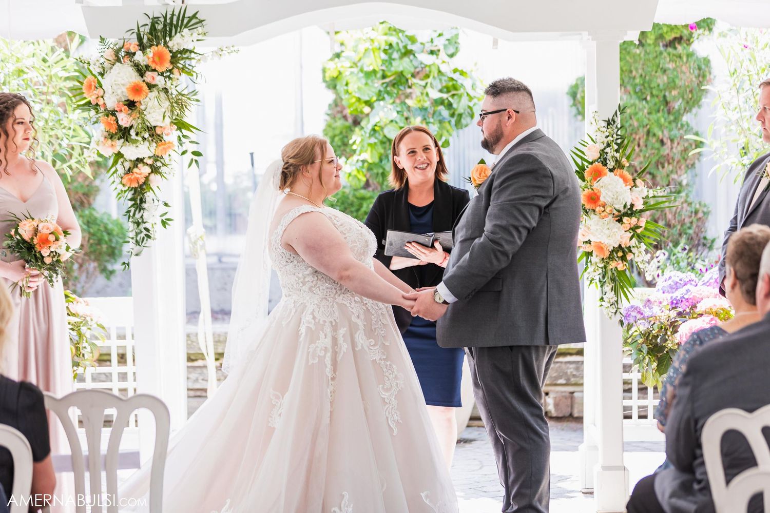 An outdoor wedding ceremony under a white tent with floral arrangements and decorative greenery.