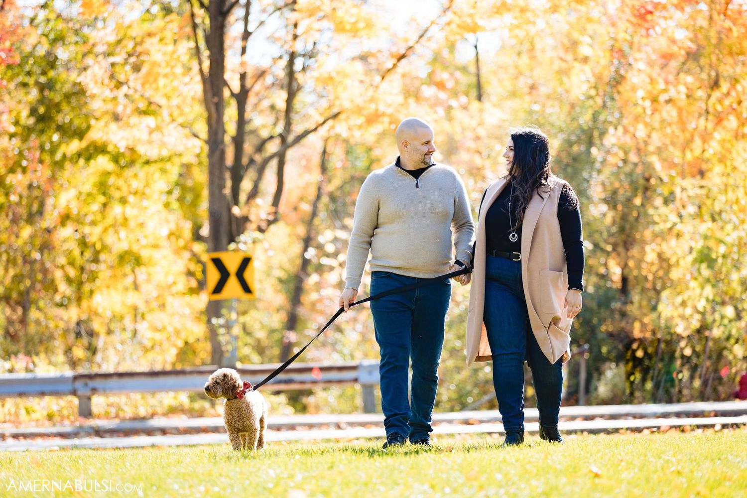 Couple walks dog on leash along autumn path with golden leaves.