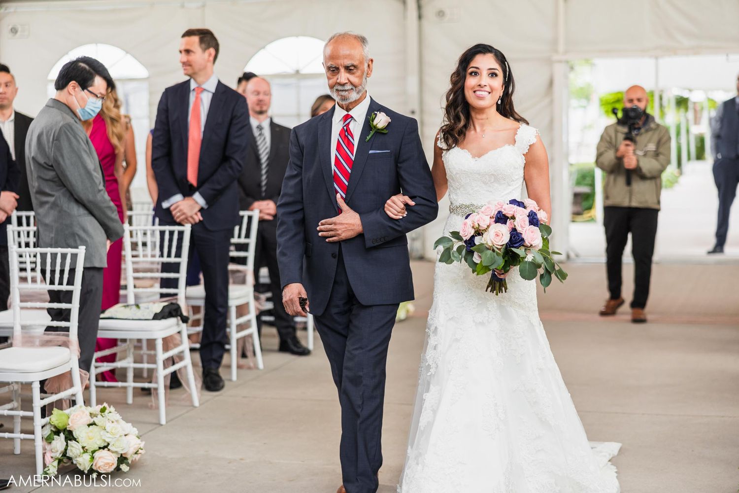 A bride walks down the aisle in a white lace dress carrying a pink and white bouquet.