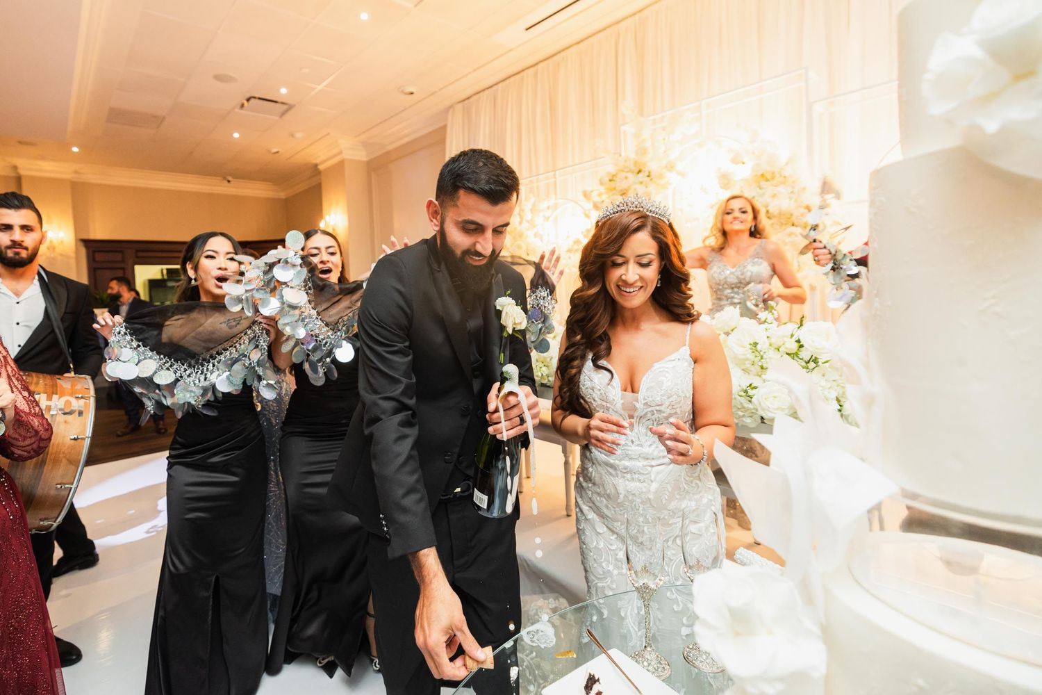 Wedding reception celebration with champagne popping and decorative white cake display.