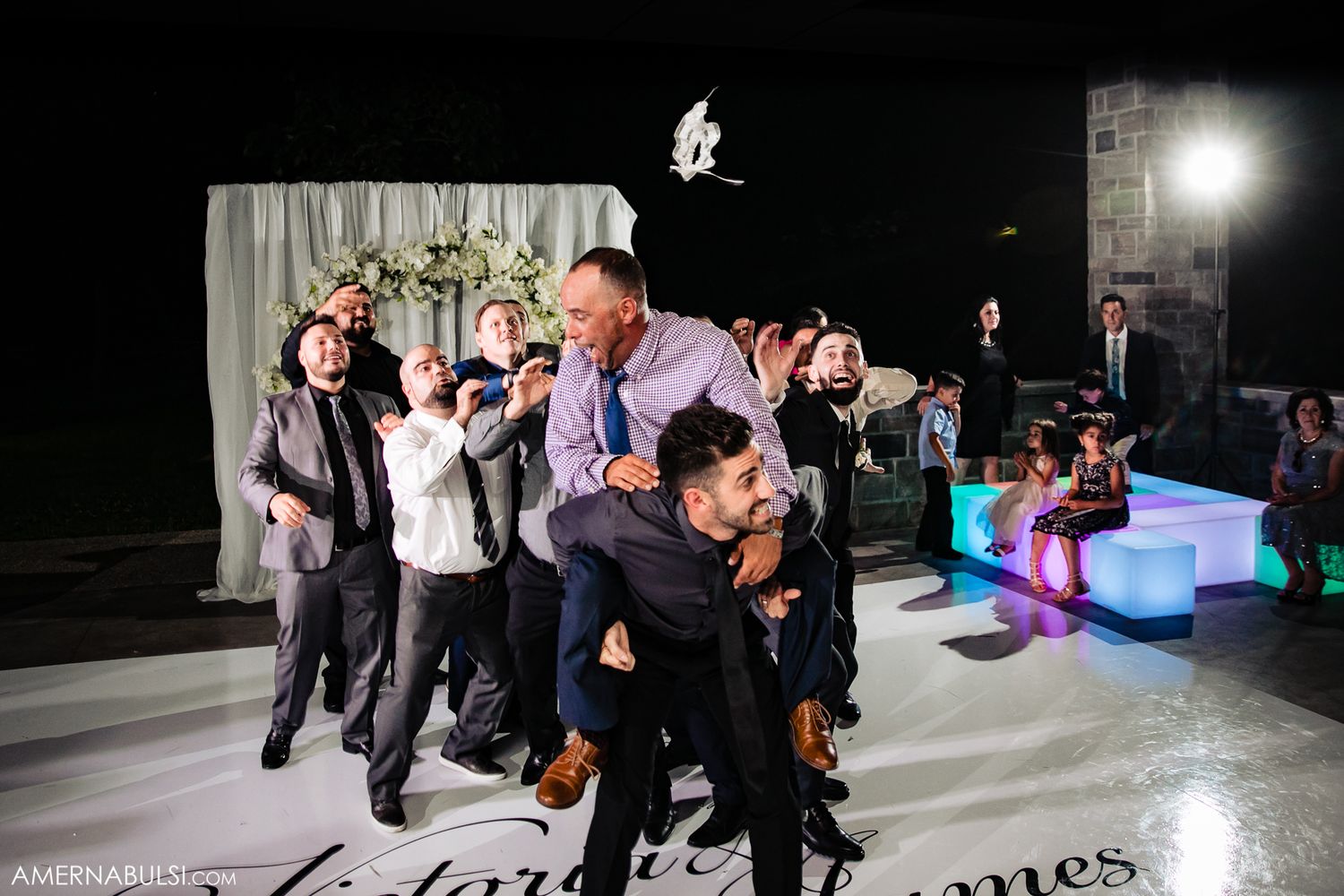 Group of wedding guests dancing energetically on illuminated dance floor at night.