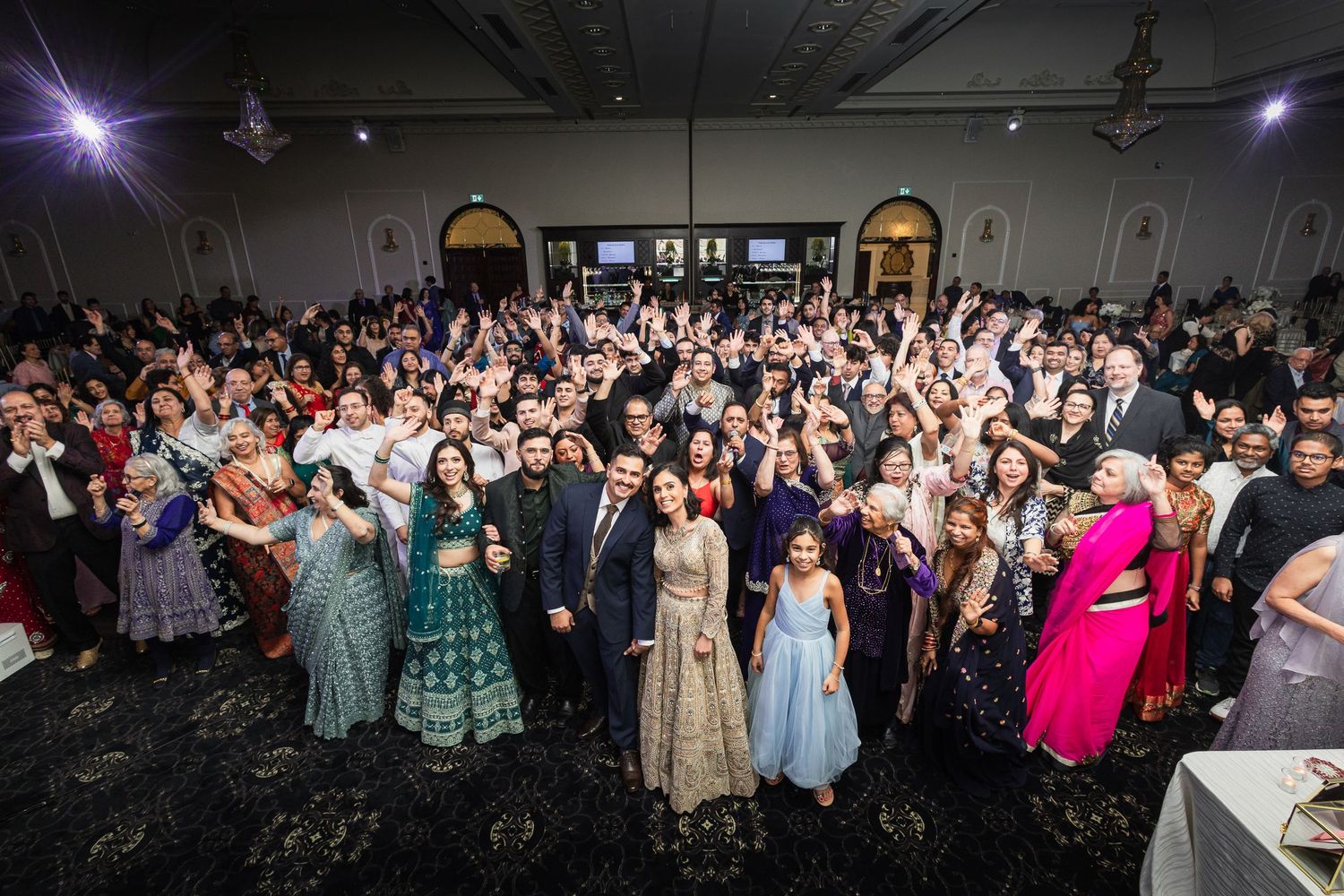 Large crowd of wedding guests dancing and celebrating in a spacious reception venue with dramatic lighting.