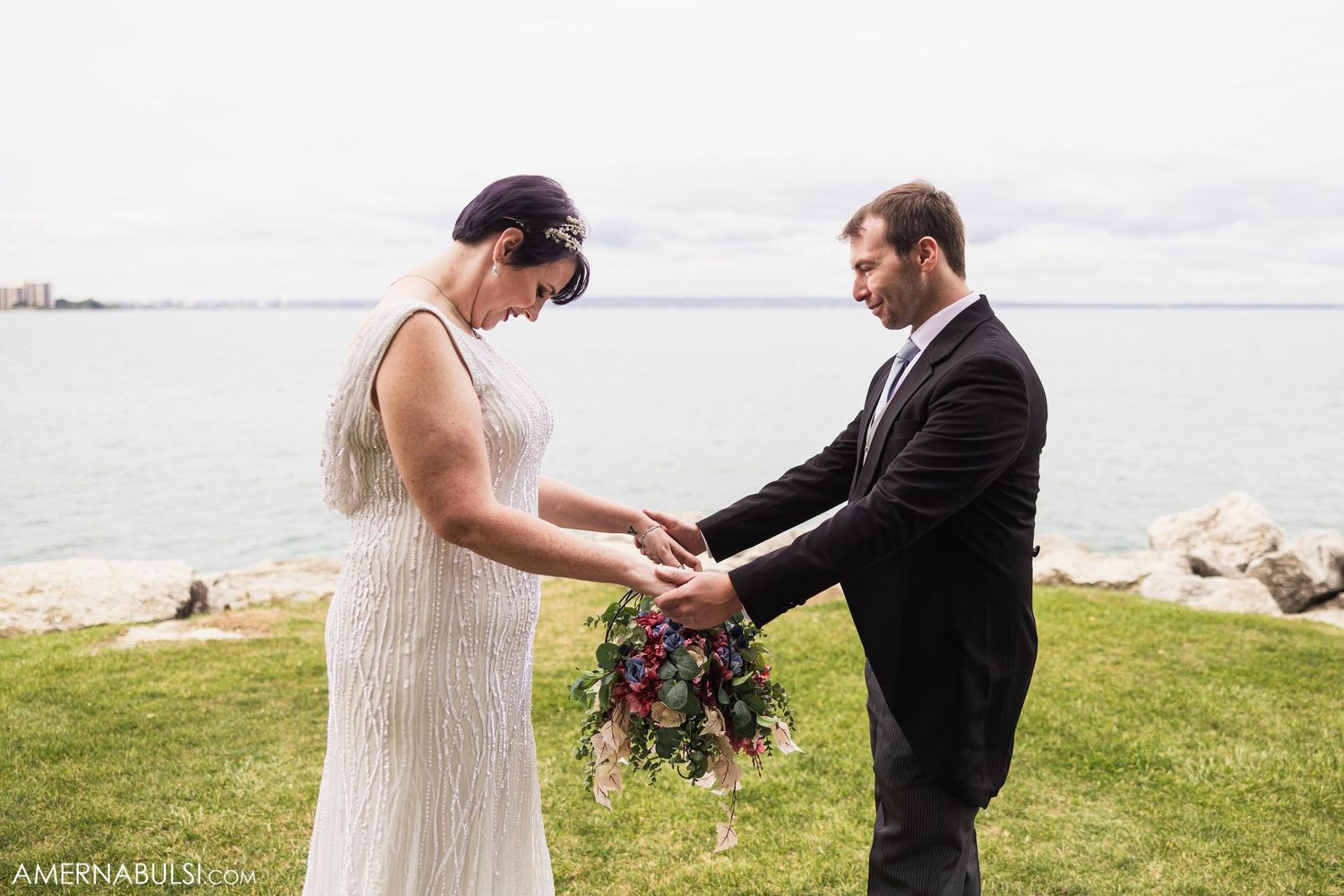 Couple shares an intimate first look moment with bouquet on a grassy waterfront setting during their wedding ceremony.