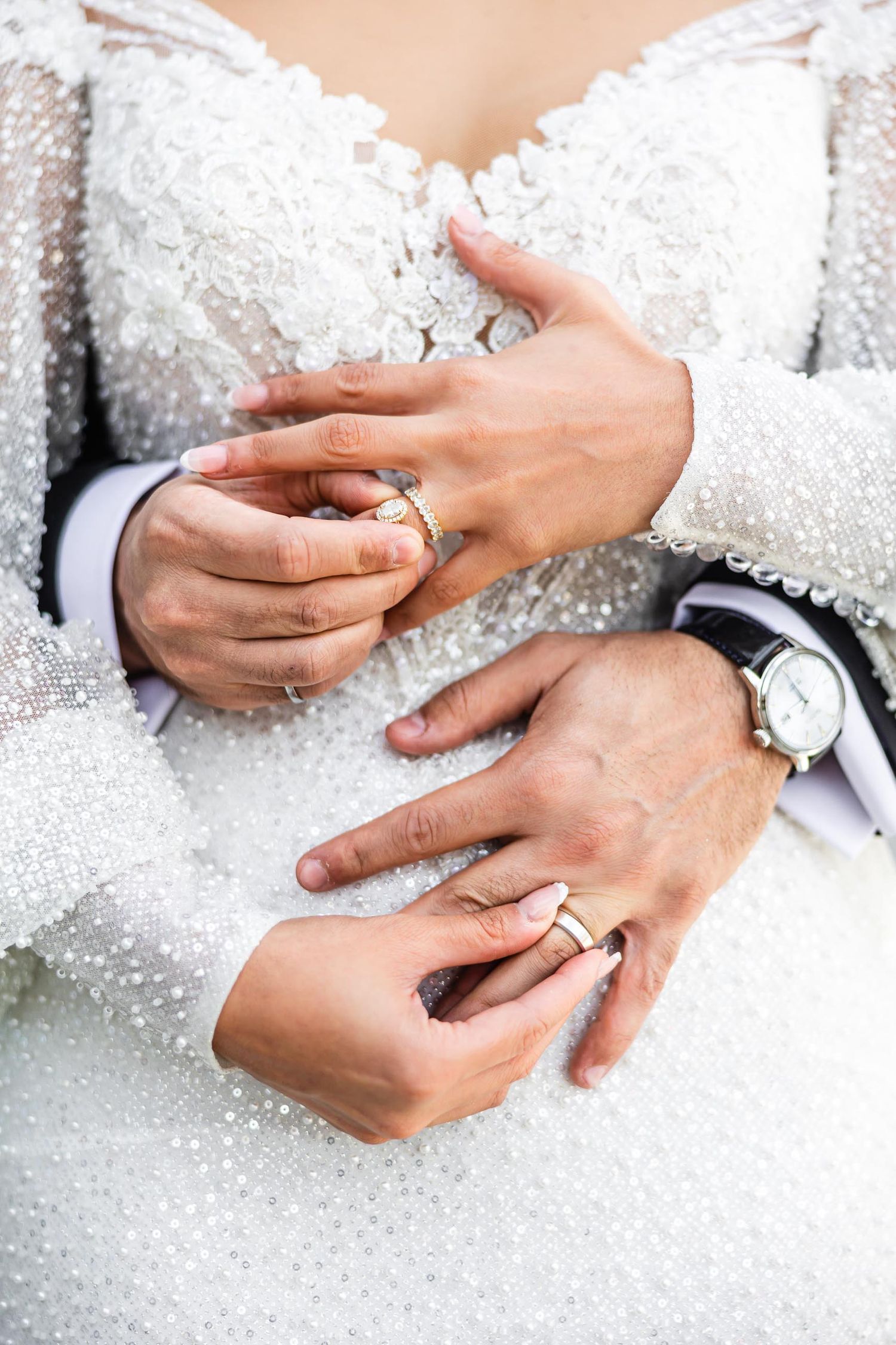 Close-up of hands displaying wedding rings against intricate beaded white dress fabric.
