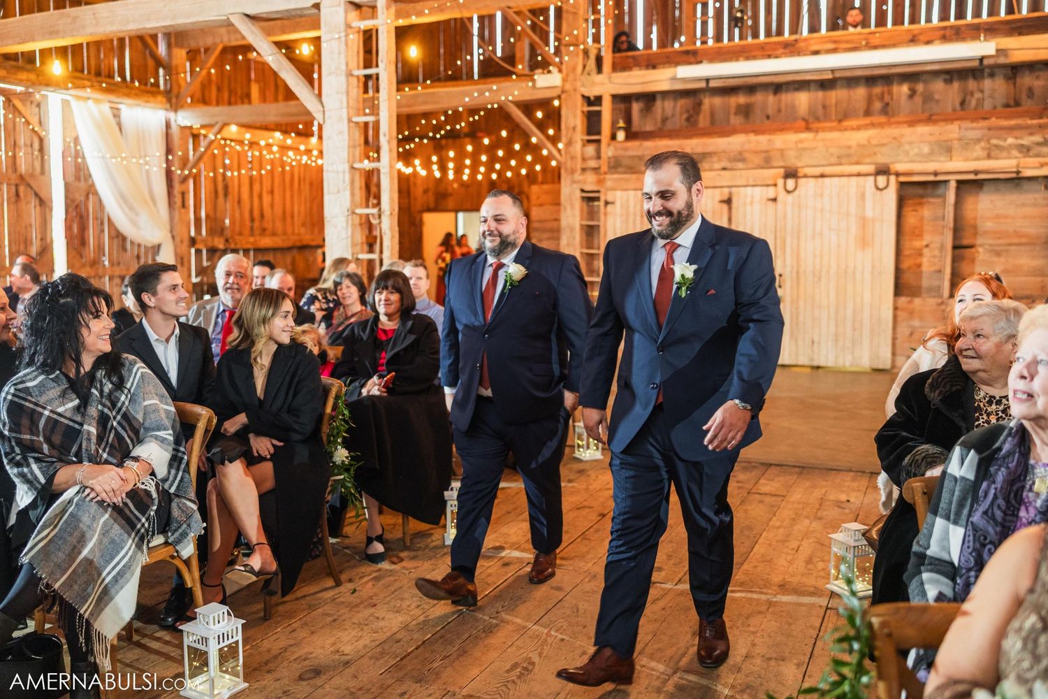 Groomsmen in navy suits with red accents walk down rustic barn venue aisle during ceremony.
