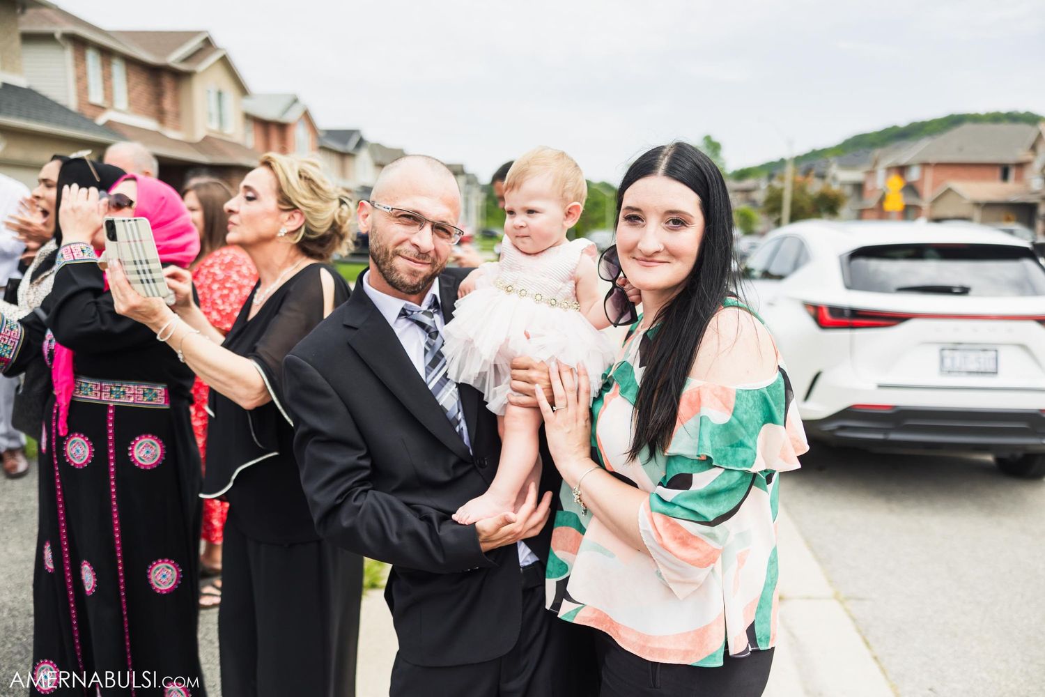 Wedding guests gather for outdoor celebration with toddler in suburban setting.