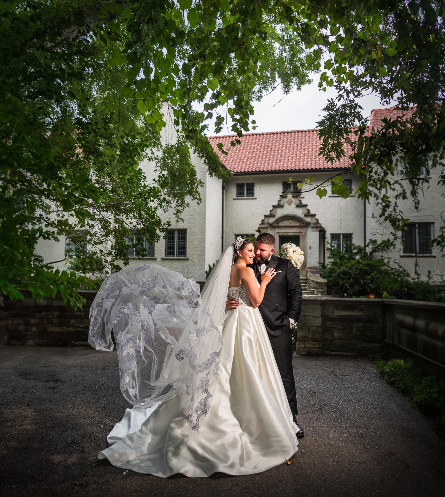 A romantic wedding photo with flowing veil in front of a white building framed by green trees.