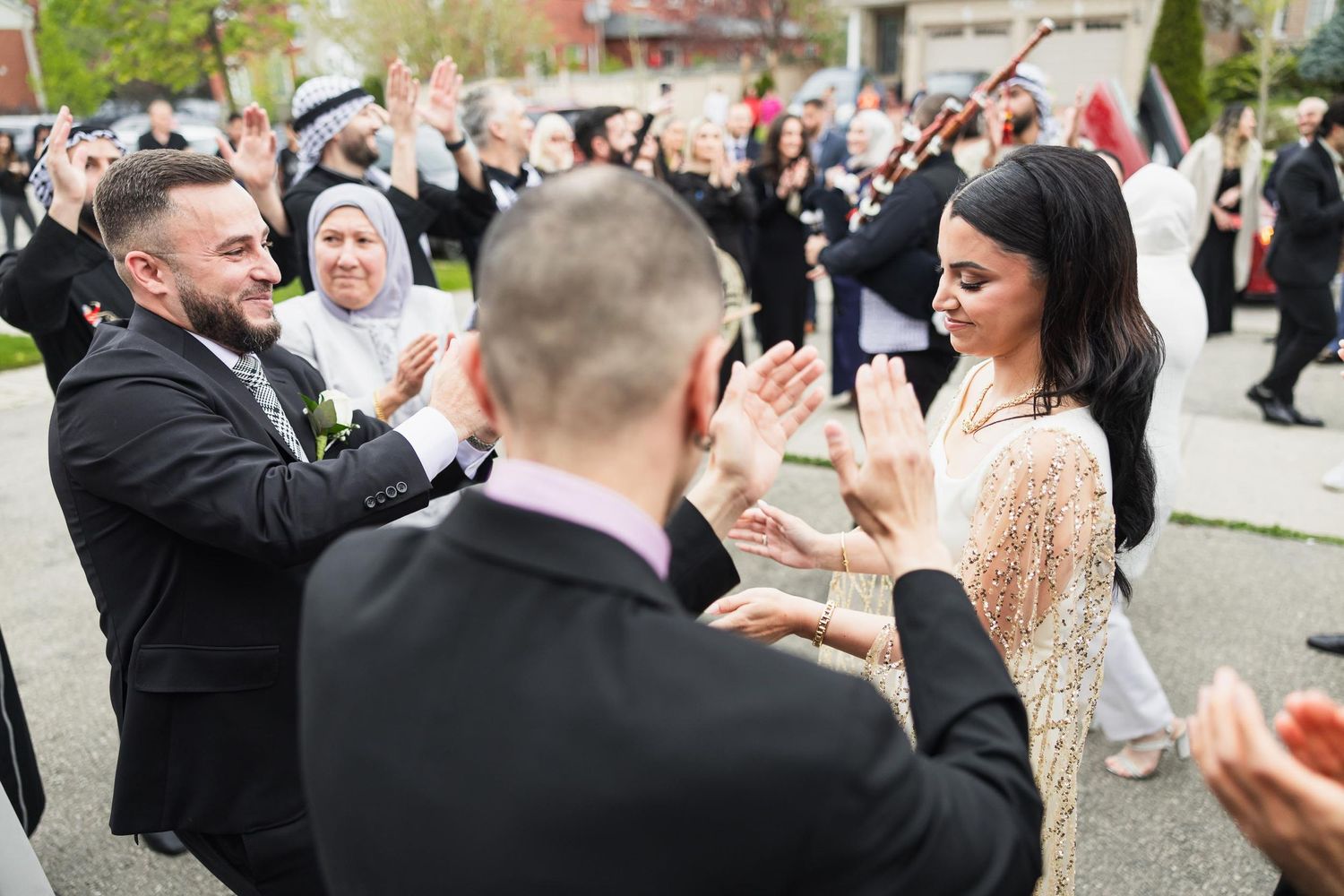 Wedding guests celebrate with dancing and applause in front of a residential building.