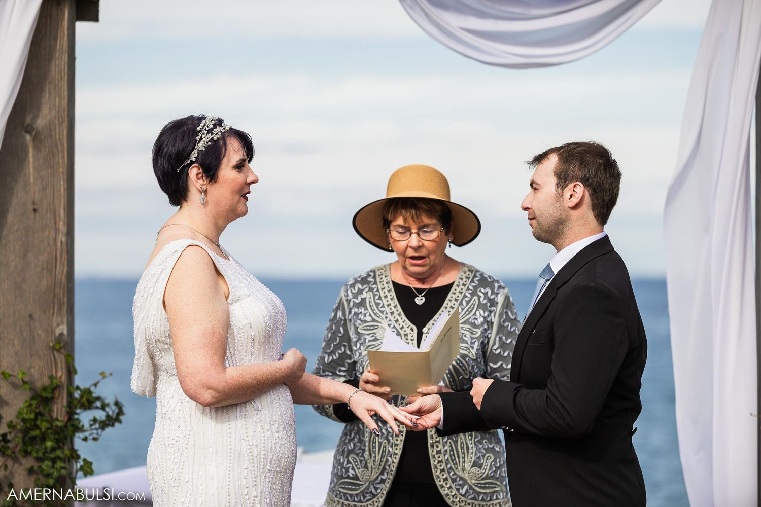 Intimate oceanside wedding ceremony with officiant wearing distinctive straw hat.