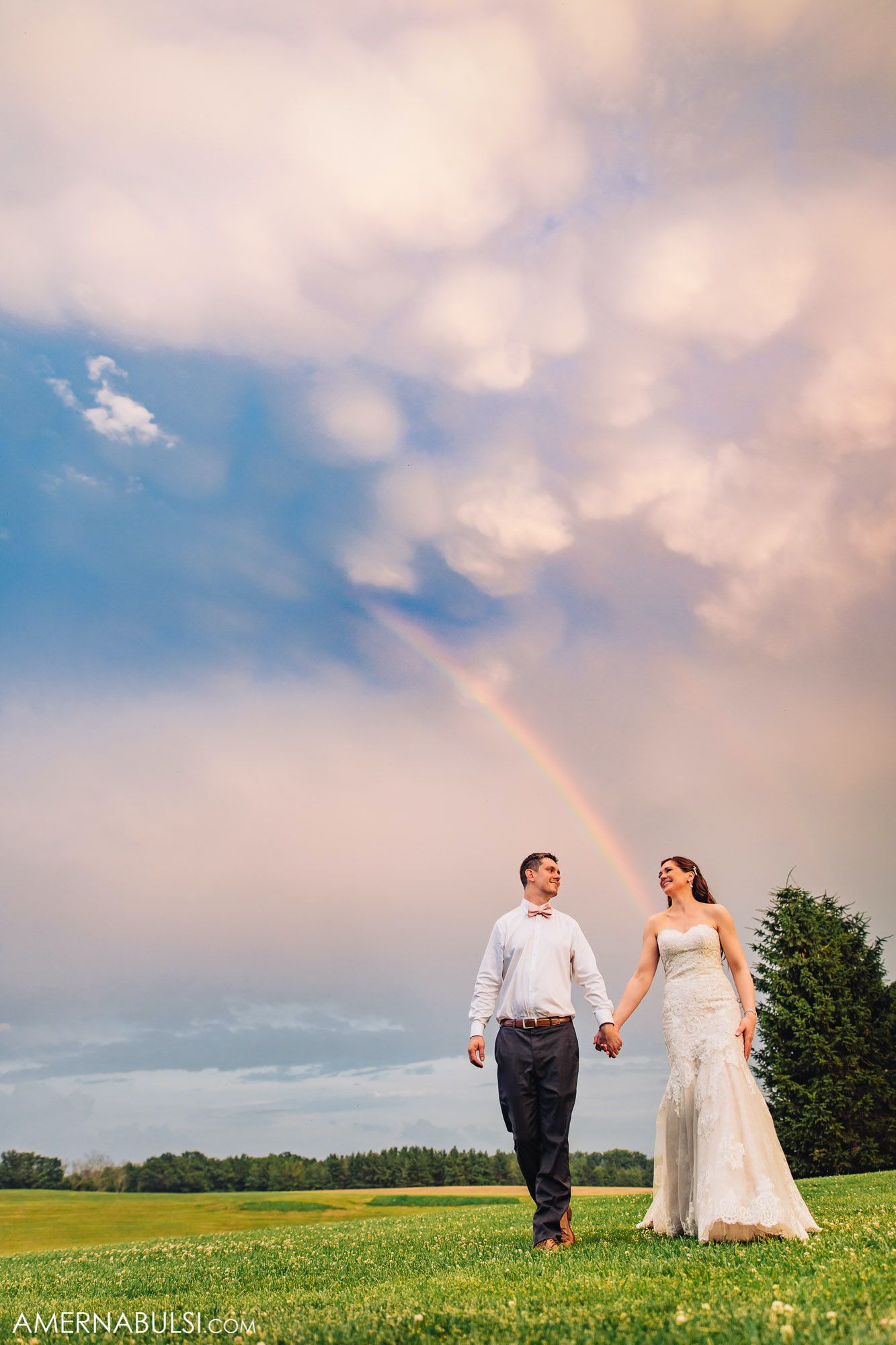 A couple walks hand in hand across a green field with a beautiful rainbow and dramatic clouds in the sky.