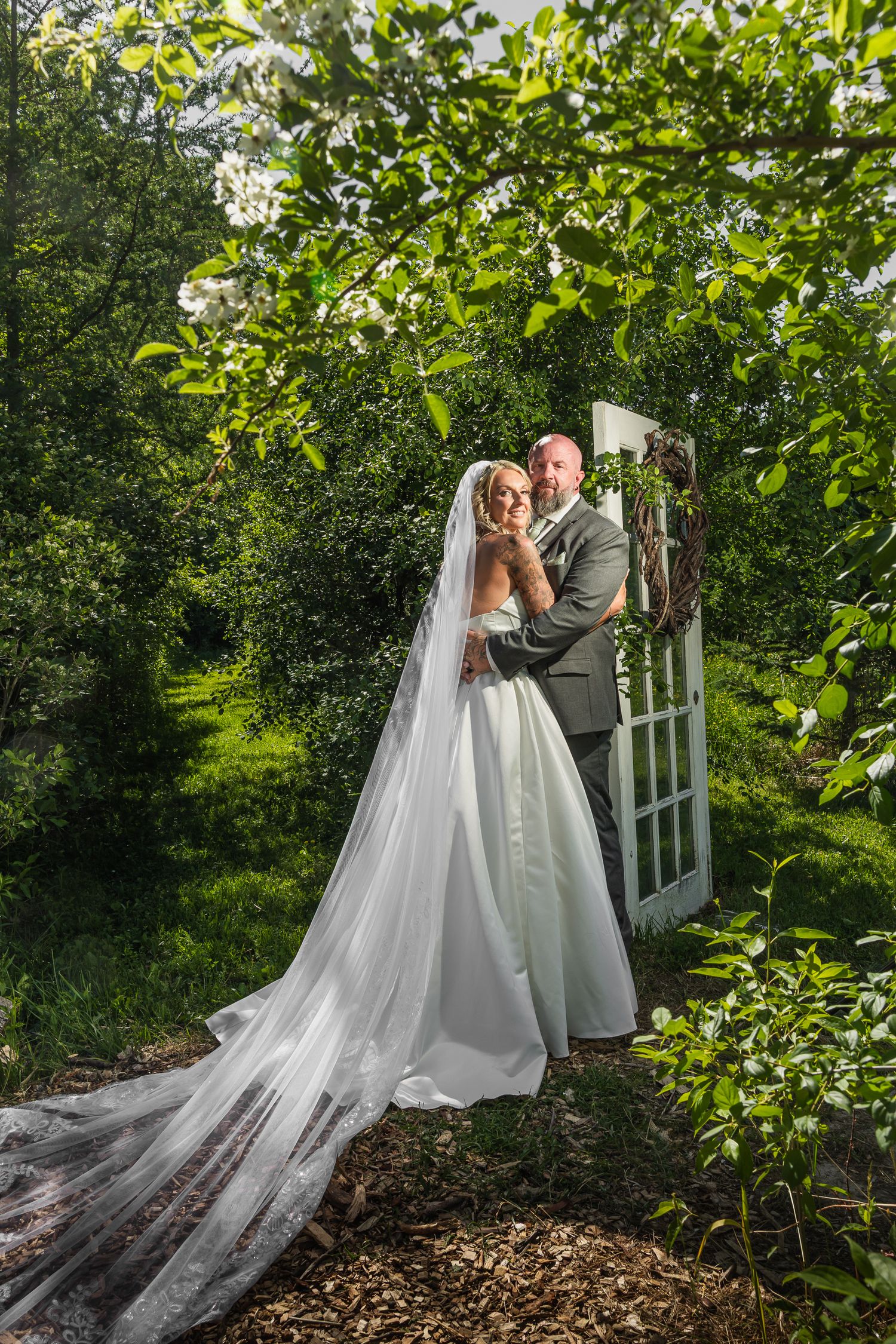 Bride and groom pose in lush garden beside rustic door arch, bride wearing long veil and white gown.