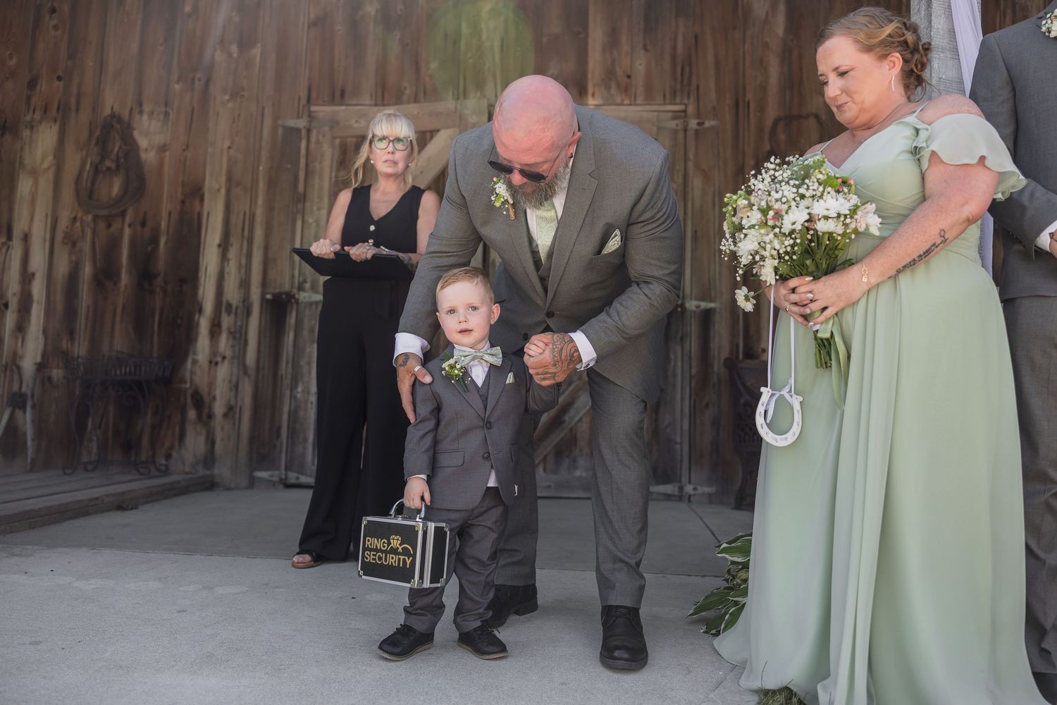 Bald man in gray suit bends to greet young ring bearer at rustic barn wedding ceremony.