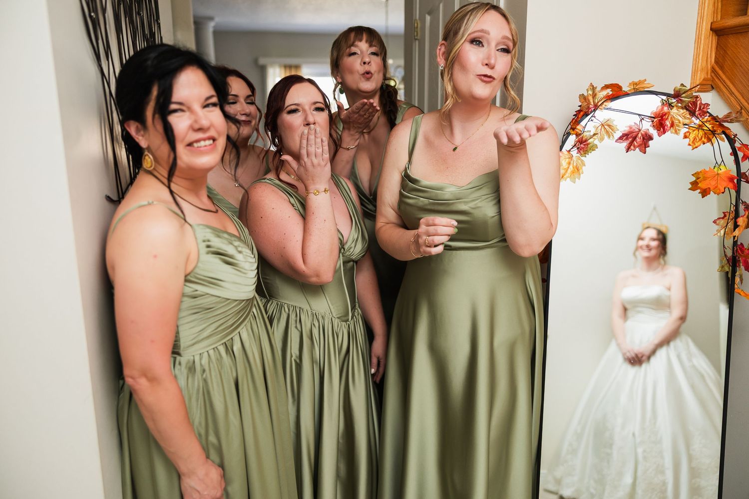 Four bridesmaids in sage green satin dresses blow kisses beside a mirror reflecting the bride in white.