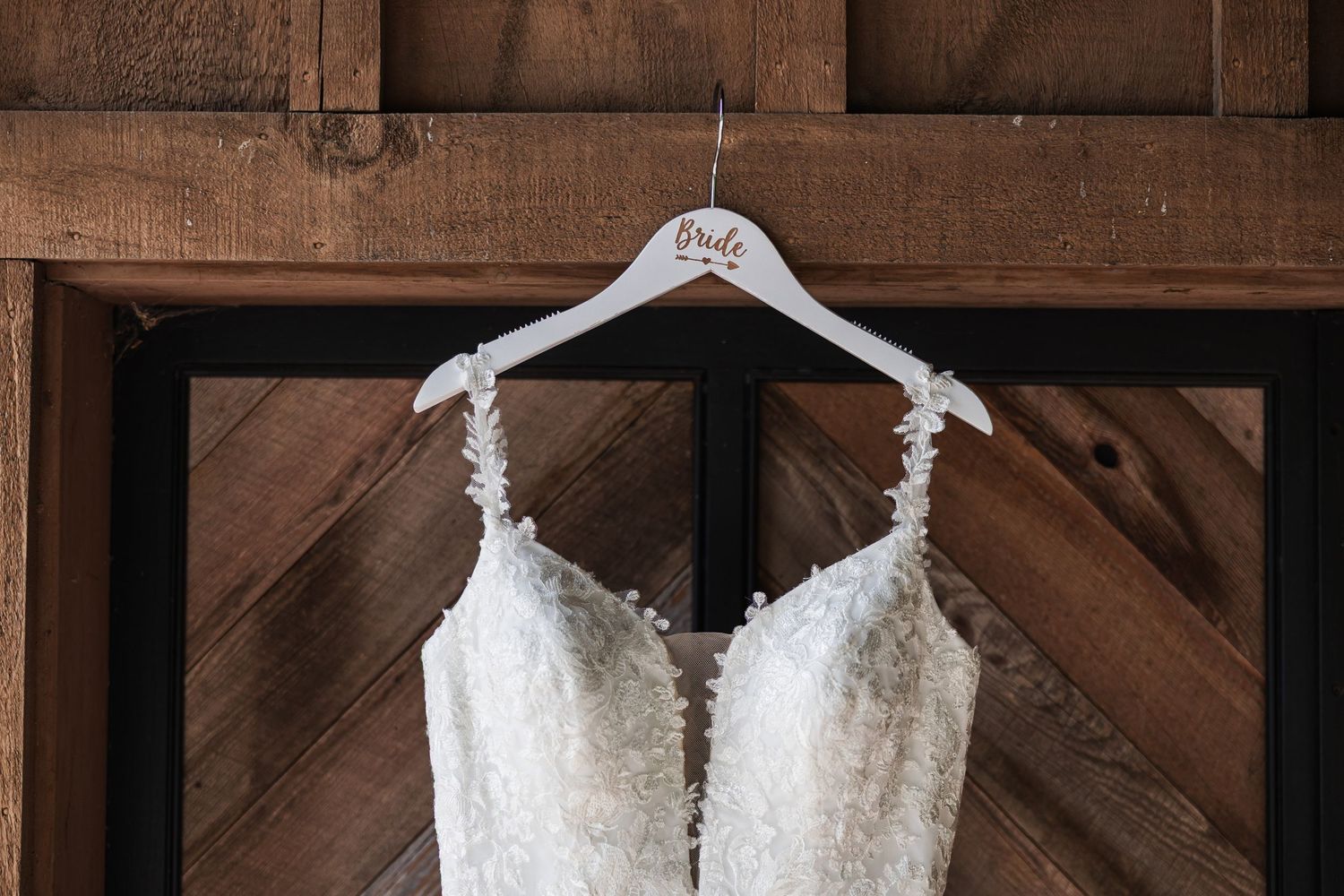 A white lace wedding dress hangs on a personalized Bride hanger against a rustic wooden barn door.