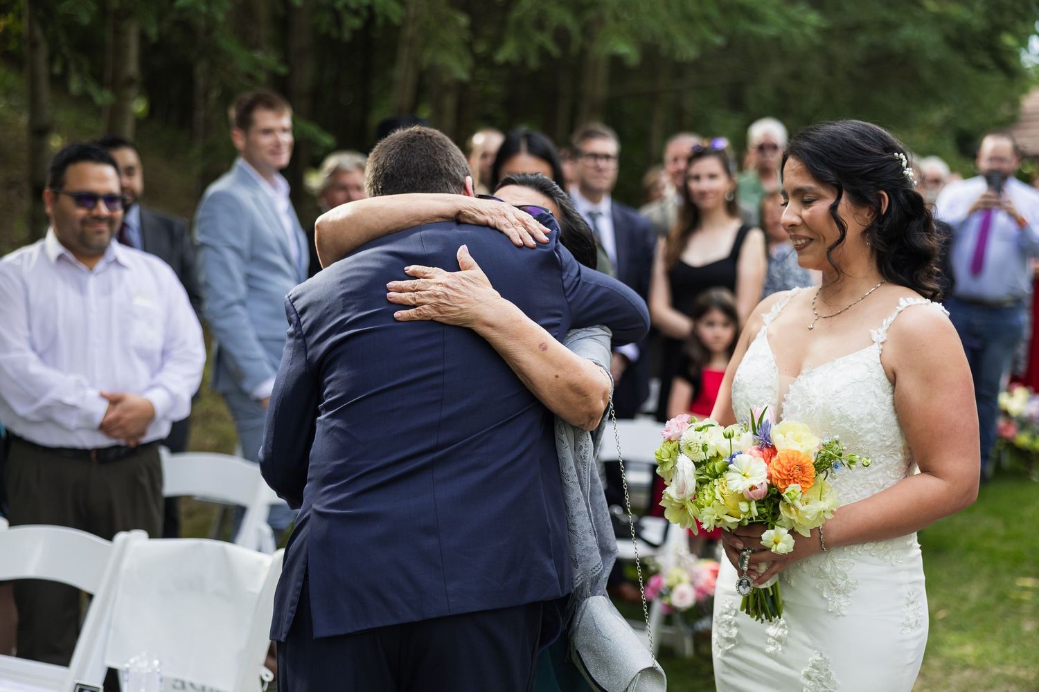 Groom embraces a guest emotionally as bride holds bouquet and smiles during outdoor wedding ceremony.