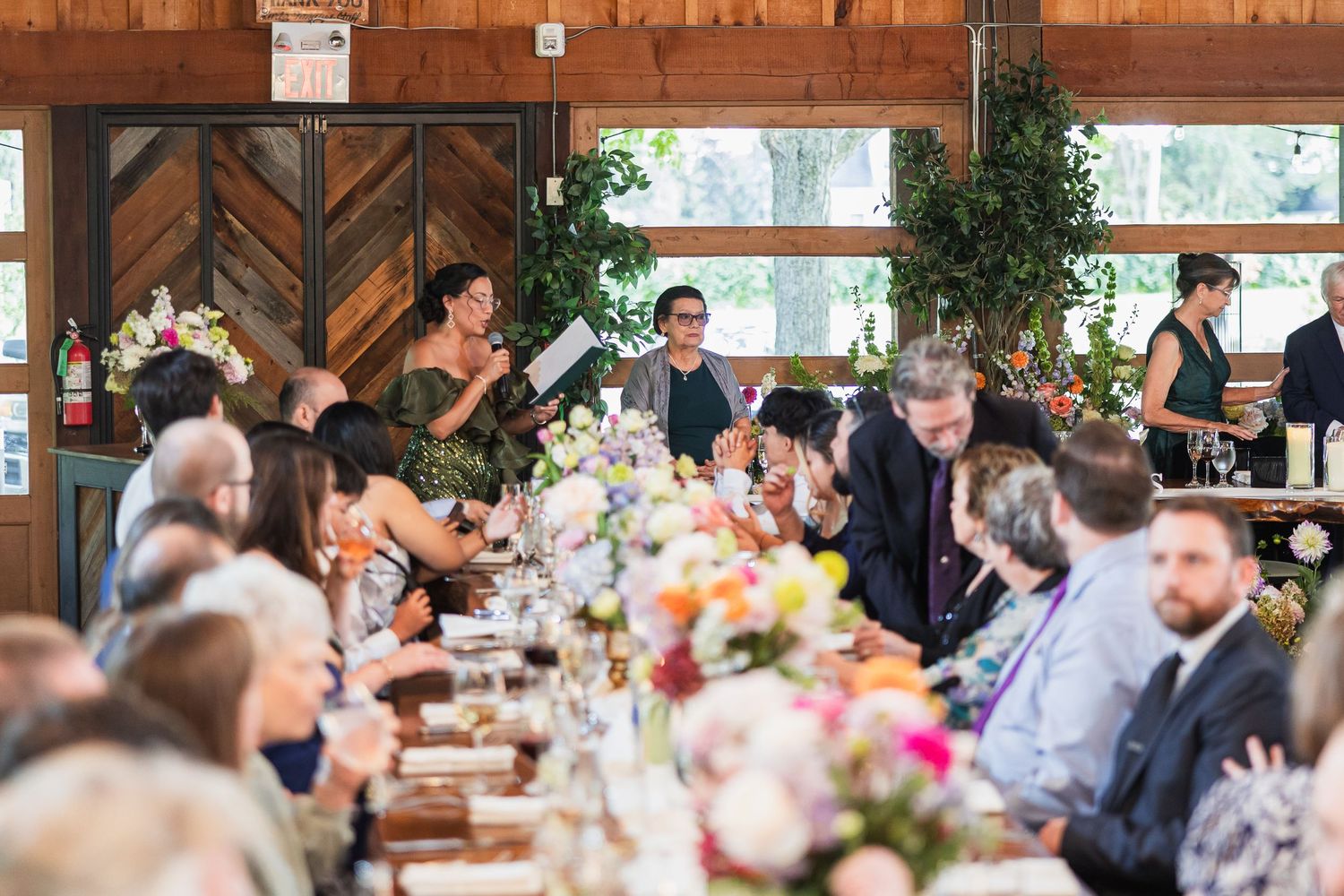 Wedding guest gives a speech at a long decorated reception table with colorful floral arrangements.