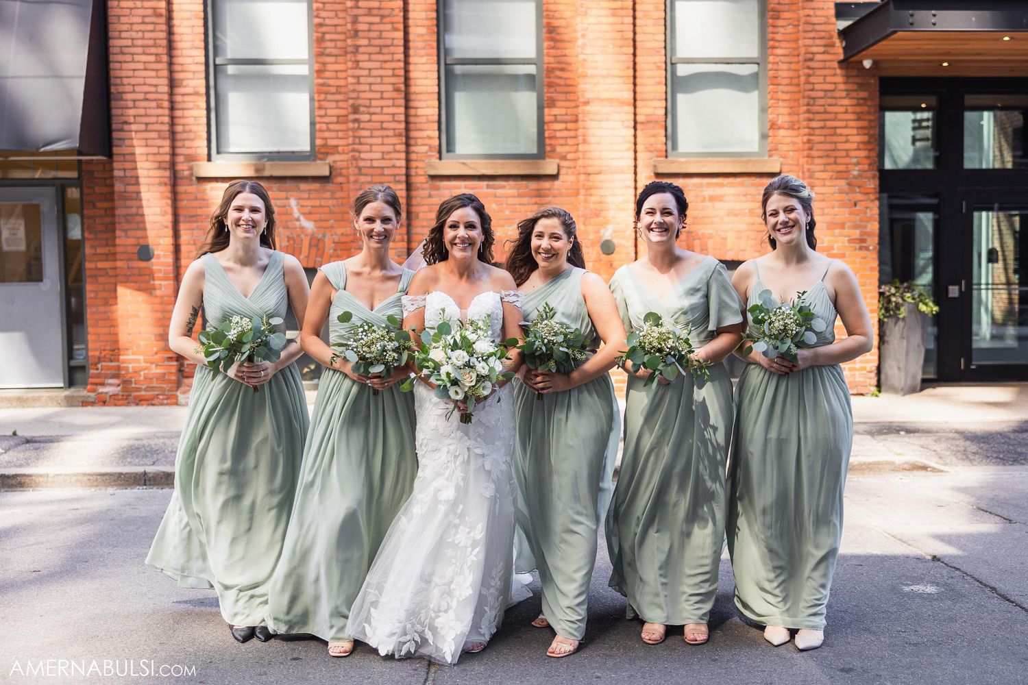 Wedding party in sage green dresses poses in front of a brick building.
