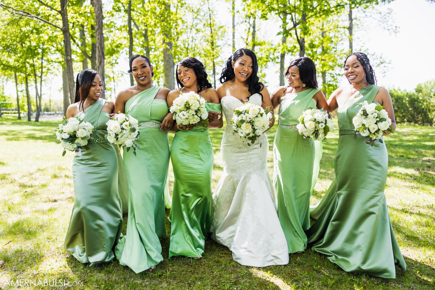 Bridal party in matching mint green dresses poses with the bride holding white bouquets outdoors.