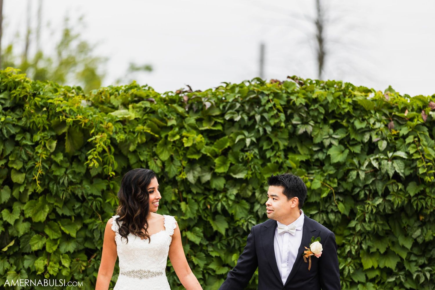 Wedding couple sits casually on lawn next to lush green hedge during outdoor photo session.