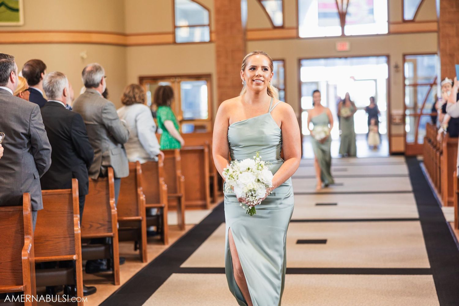 A woman in a strapless mint gown walks gracefully down the church aisle carrying white flowers.