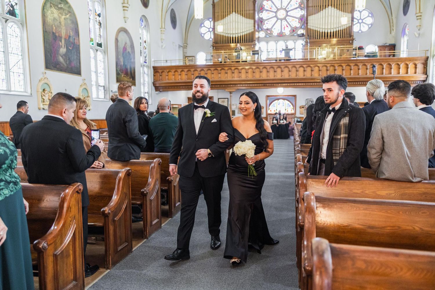 Wedding party members walk between wooden pews in ornate church interior.