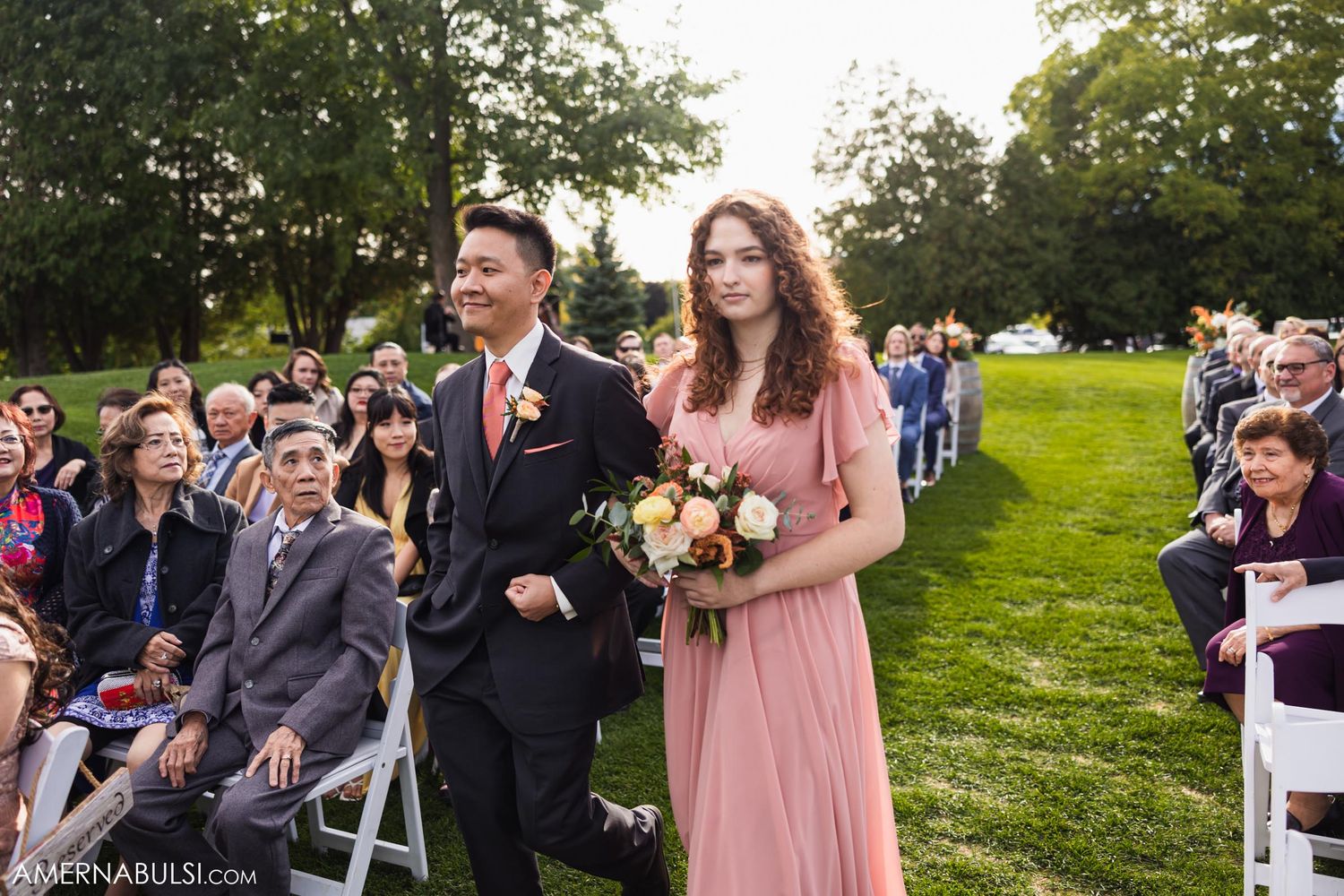 A bridesmaid and groomsman process down a grassy aisle lined with seated wedding guests.