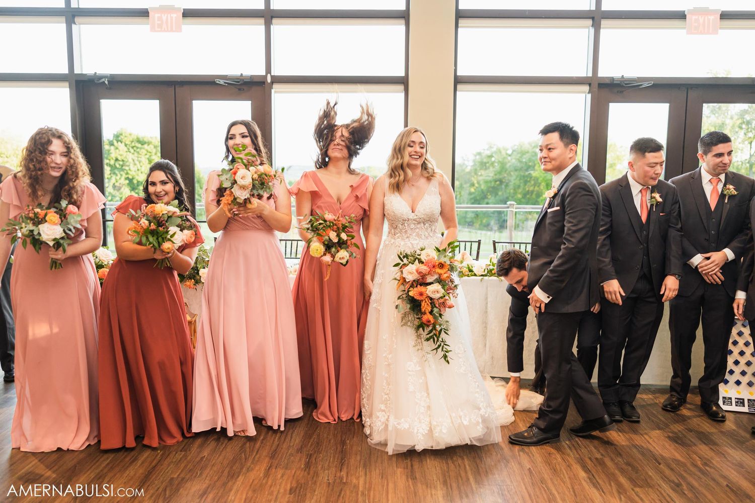 Wedding party in pink bridesmaid dresses and black suits pose together in front of large windows.