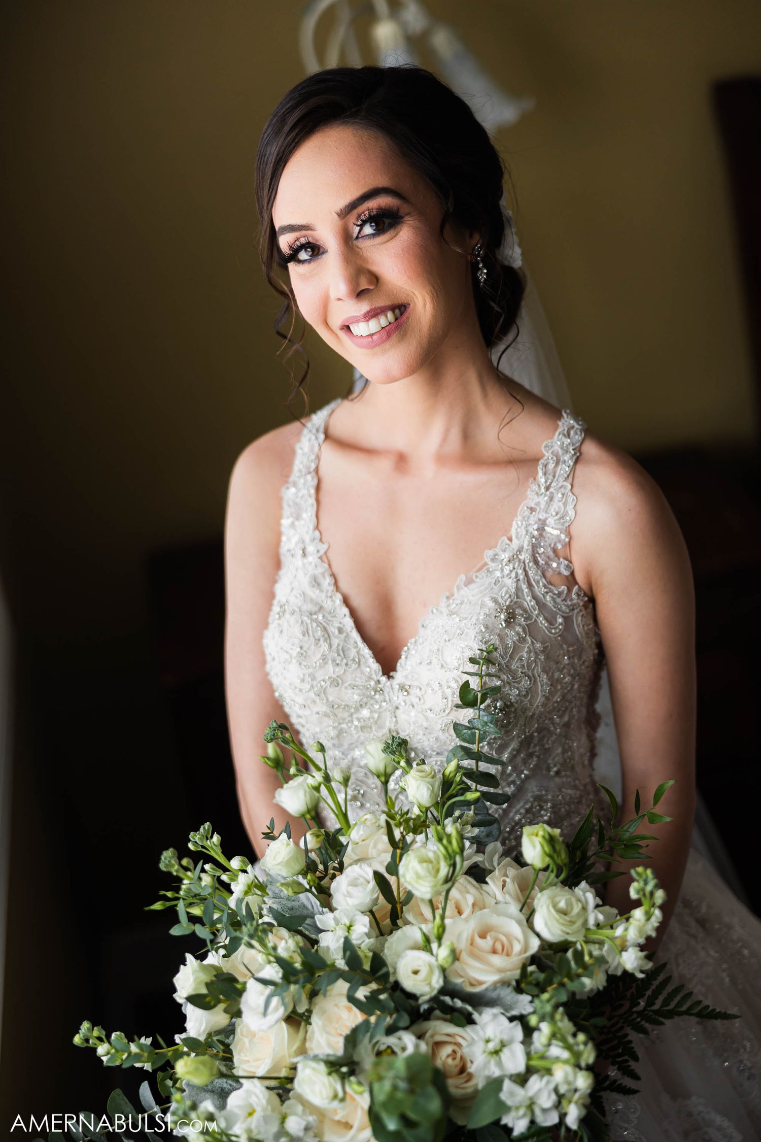 An elegant bridal portrait with a lush white and green bouquet against a warm yellow wall.
