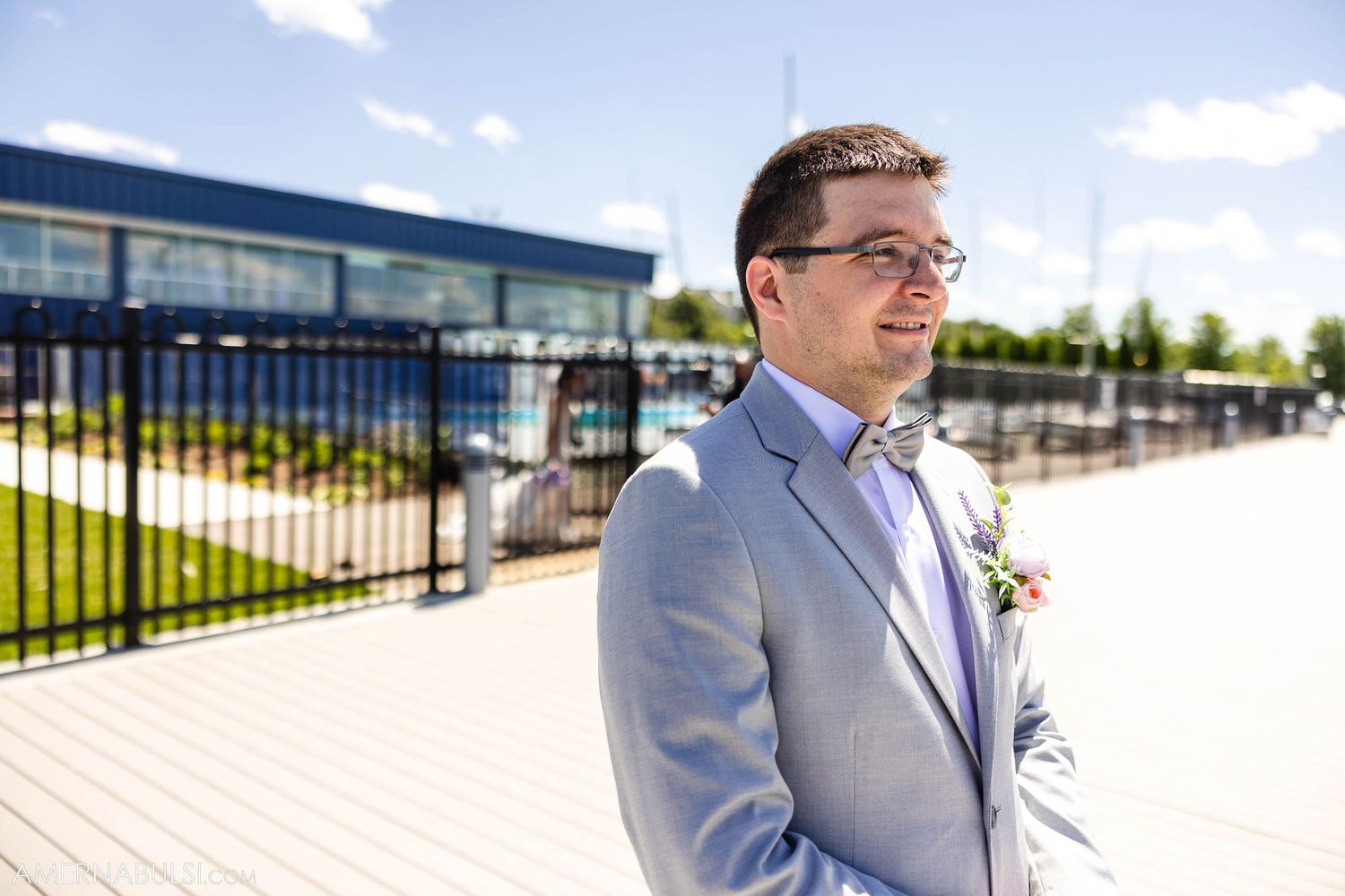 Groom in light gray suit stands in sunlight near modern building.