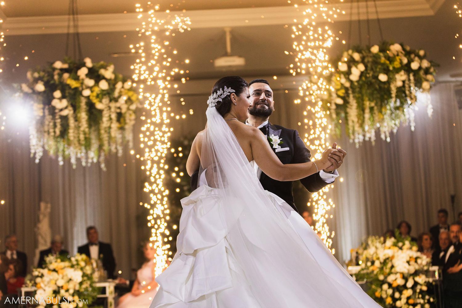 Newlyweds share their first dance under stunning cascading lights and floral decorations at an elegant wedding reception.