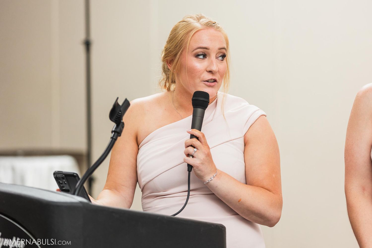 Wedding guest in a one-shoulder dress gives a speech at the reception with microphone in hand.