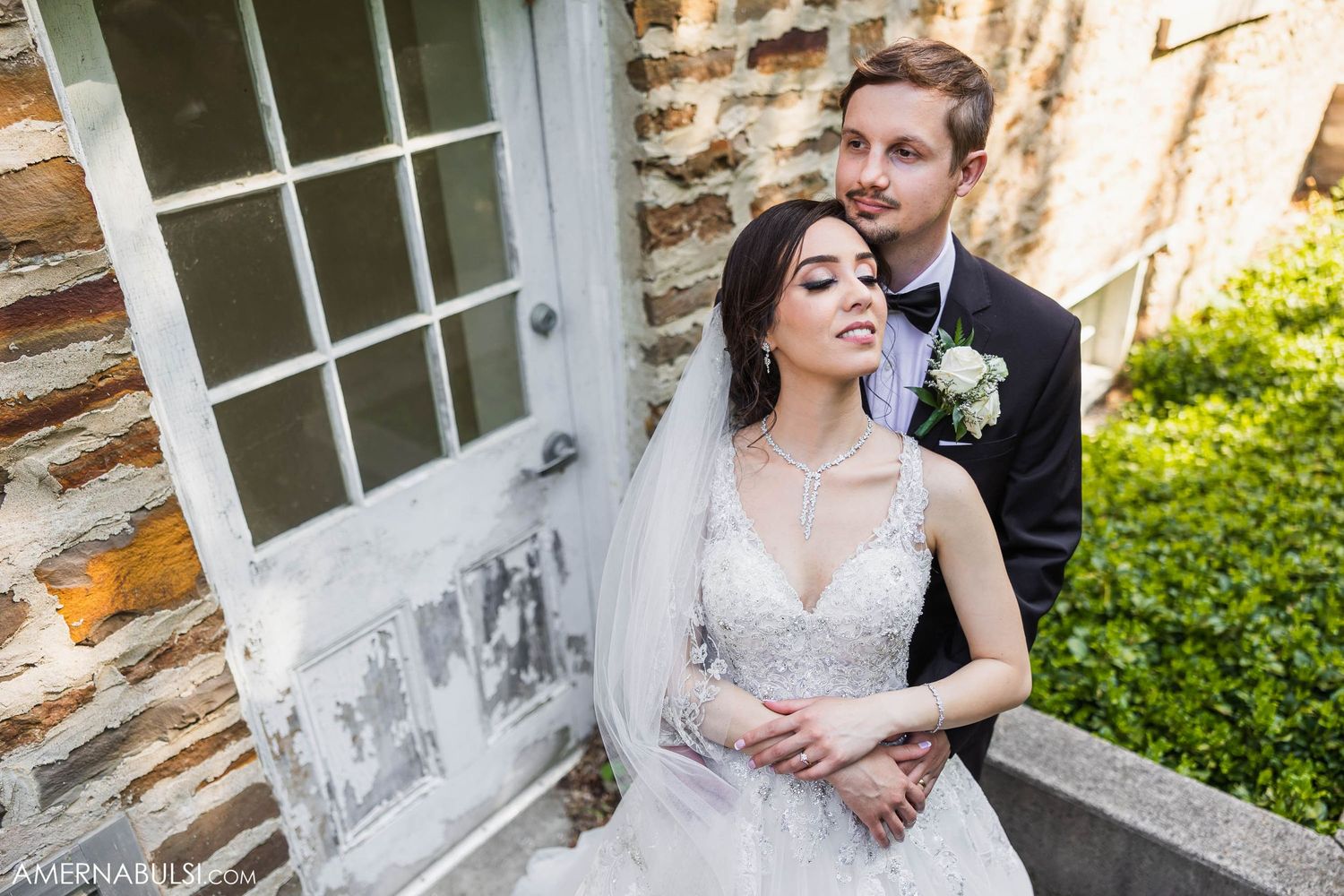 Elegant wedding couple poses by stone wall in formal attire with flowing veil.