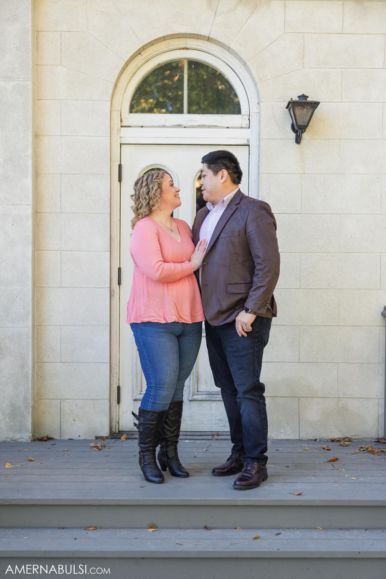A couple stands close together on a porch with an arched doorway behind them.