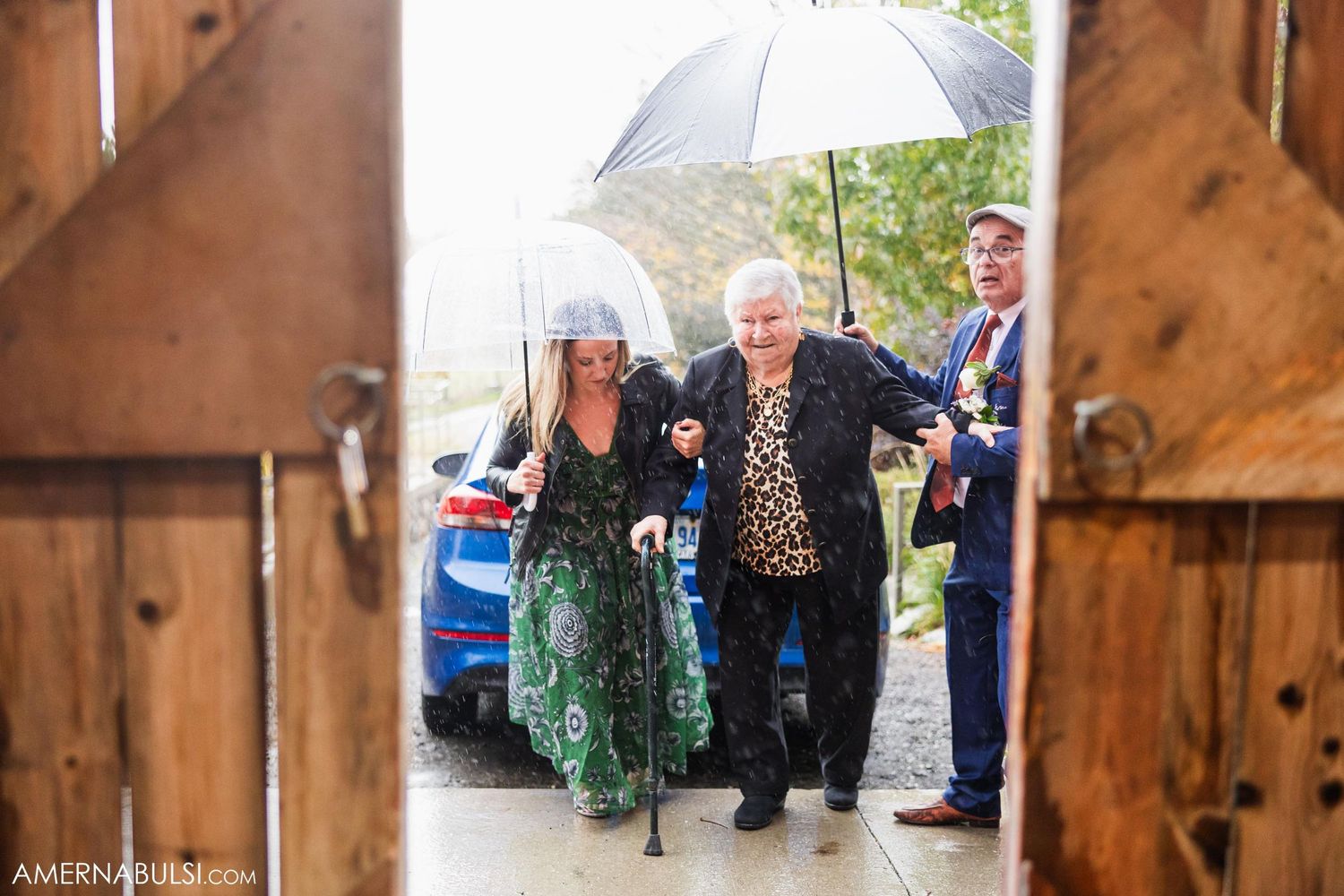 Wedding guests arrive under umbrella through rustic wooden barn doors on rainy day.