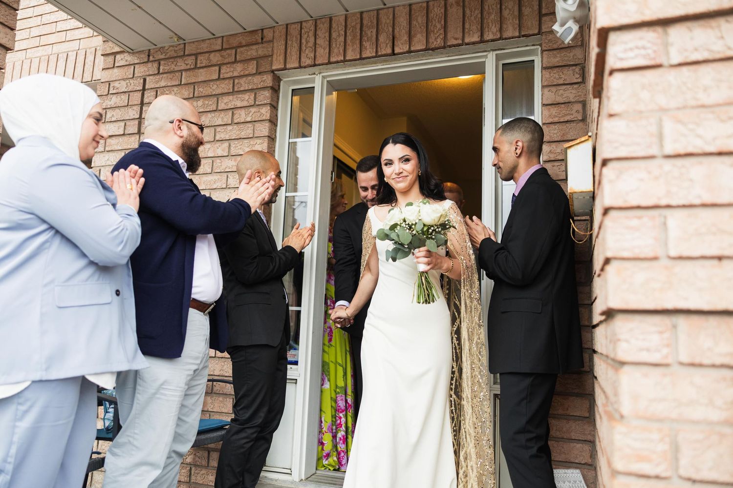 A bride in a white dress holding a bouquet stands in a doorway surrounded by well-wishers.