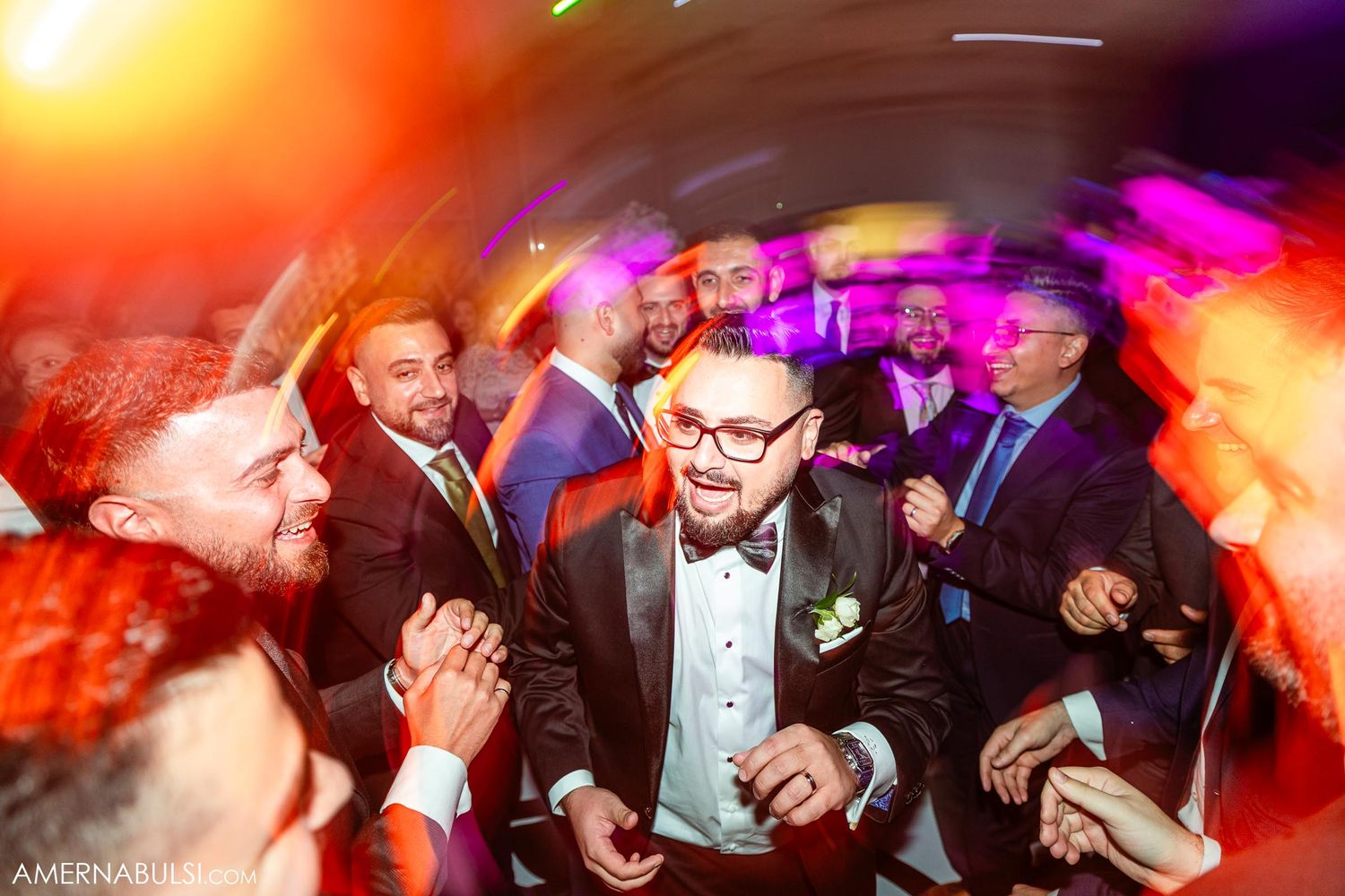 Wedding reception dance floor filled with guests in formal attire under dramatic colorful lighting.