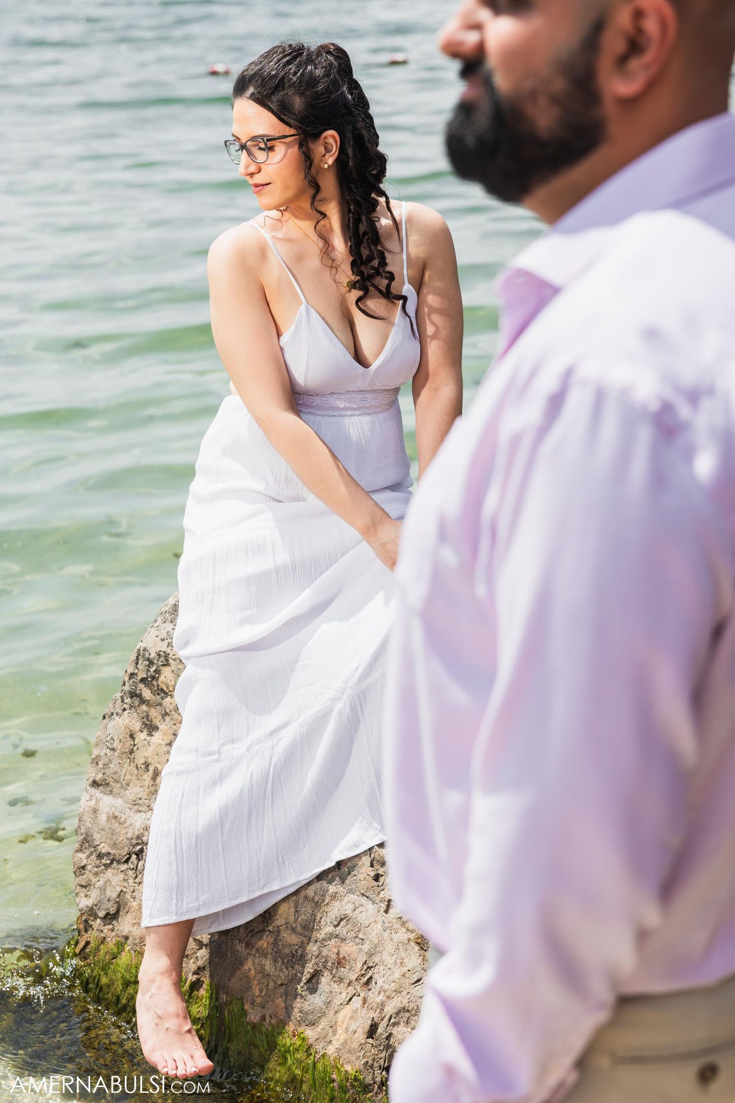 Woman in white dress stands on rocky shore while companion looks on by the waterfront.